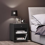 Schwarzer Nachttisch auf Rollen mit Schublade und offenen Fächern neben einem grauen Bett, darauf schwarze Tischlampe, Bilderrahmen und kleiner Wecker; Perspektive: frontale Seitenansicht auf Nachttisch und Bett.