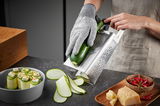 Perspektive von der Seite: Eine Vierkantreibe V-CUT wird verwendet, um eine Zucchini in dünne Scheiben zu schneiden. Eine Person trägt einen Schnittschutzhandschuh und hält die Zucchini auf der Reibe. Auf dem Tisch liegen geschnittene Zucchinischeiben, ei
