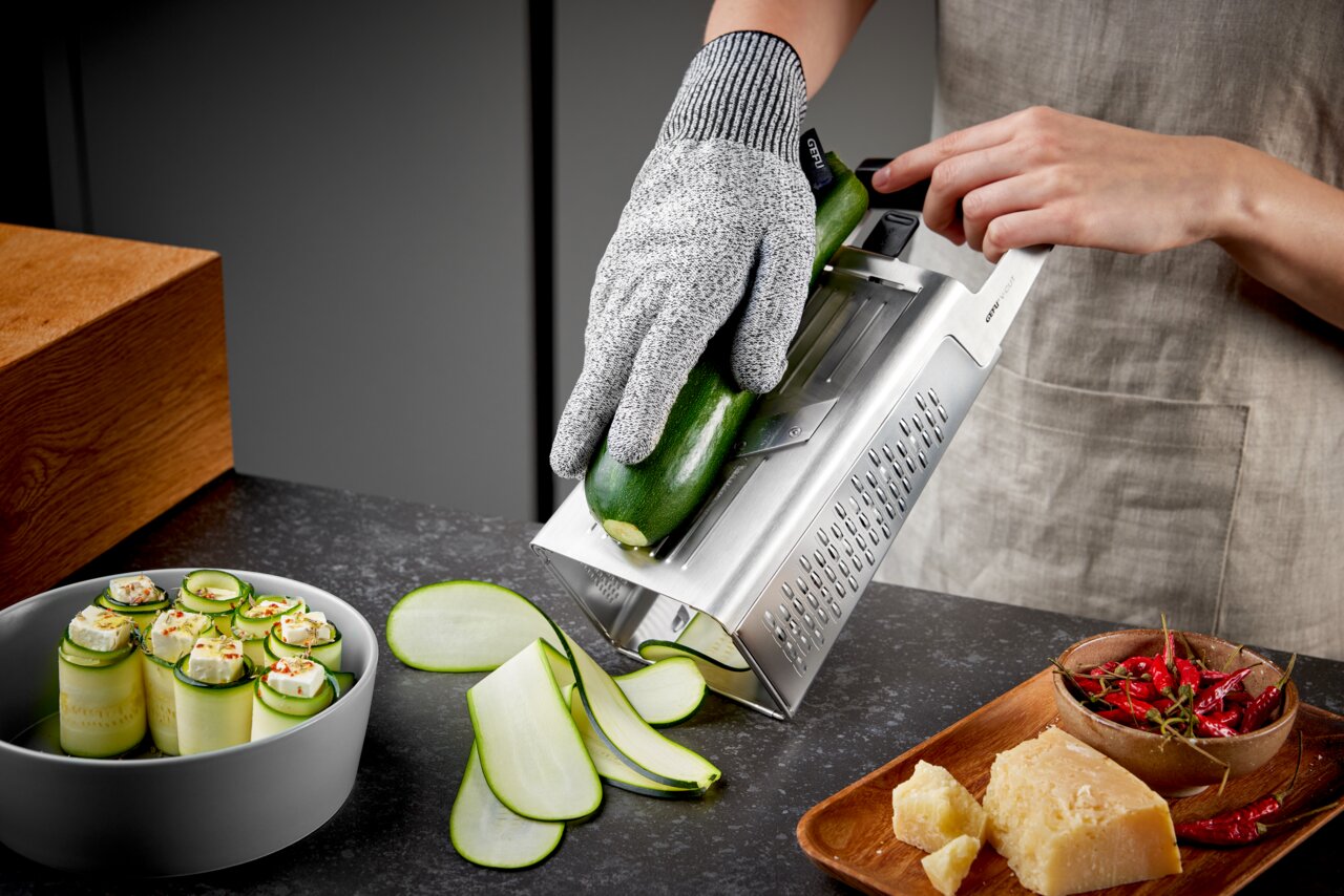 Perspektive von der Seite: Eine Vierkantreibe V-CUT wird verwendet, um eine Zucchini in dünne Scheiben zu schneiden. Eine Person trägt einen Schnittschutzhandschuh und hält die Zucchini auf der Reibe. Auf dem Tisch liegen geschnittene Zucchinischeiben, ei