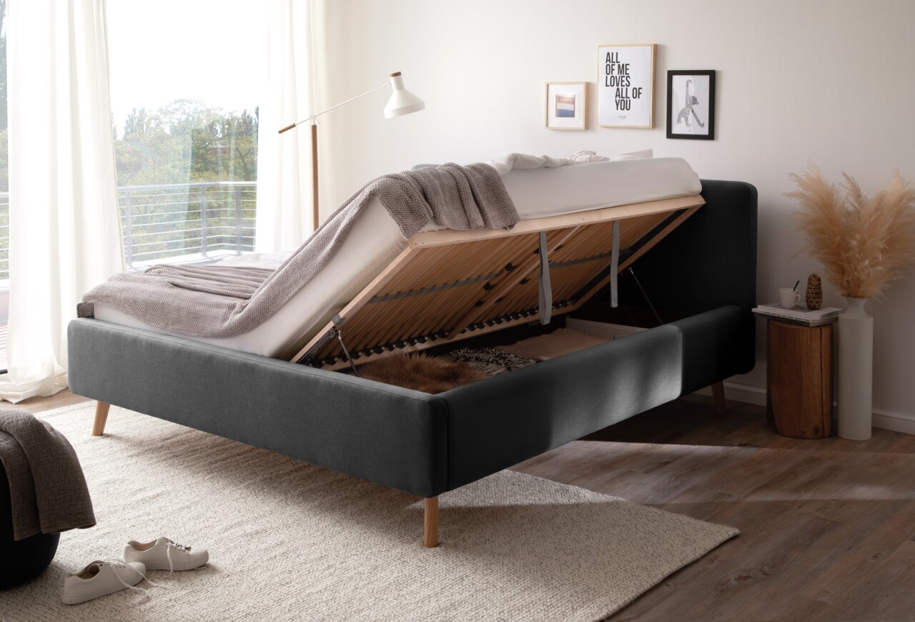 Seitliche Perspektive auf ein Polsterbett mit hochgeklappter Matratze, das Stauraum darunter zeigt. Das Bett steht in einem hellen Raum mit moderner Dekoration.