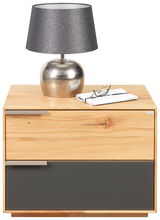 Nachtkonsole aus Holz mit zwei Schubladen, eine in Holzoptik und eine in dunklem Grau. Auf der Konsole steht eine silberne Lampe mit grauem Schirm und eine Brille auf einem Buch. Frontalansicht.