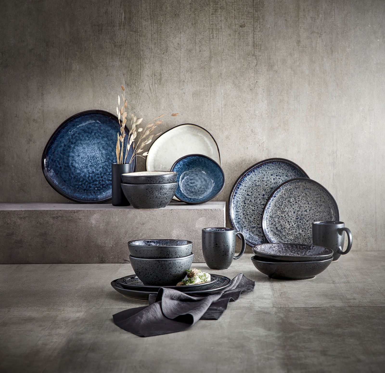 Kombiservice Sören in verschiedenen Blautönen, bestehend aus Tellern, Schalen und Tassen, auf einer strukturierten Oberfläche arrangiert. Seitliche Perspektive.