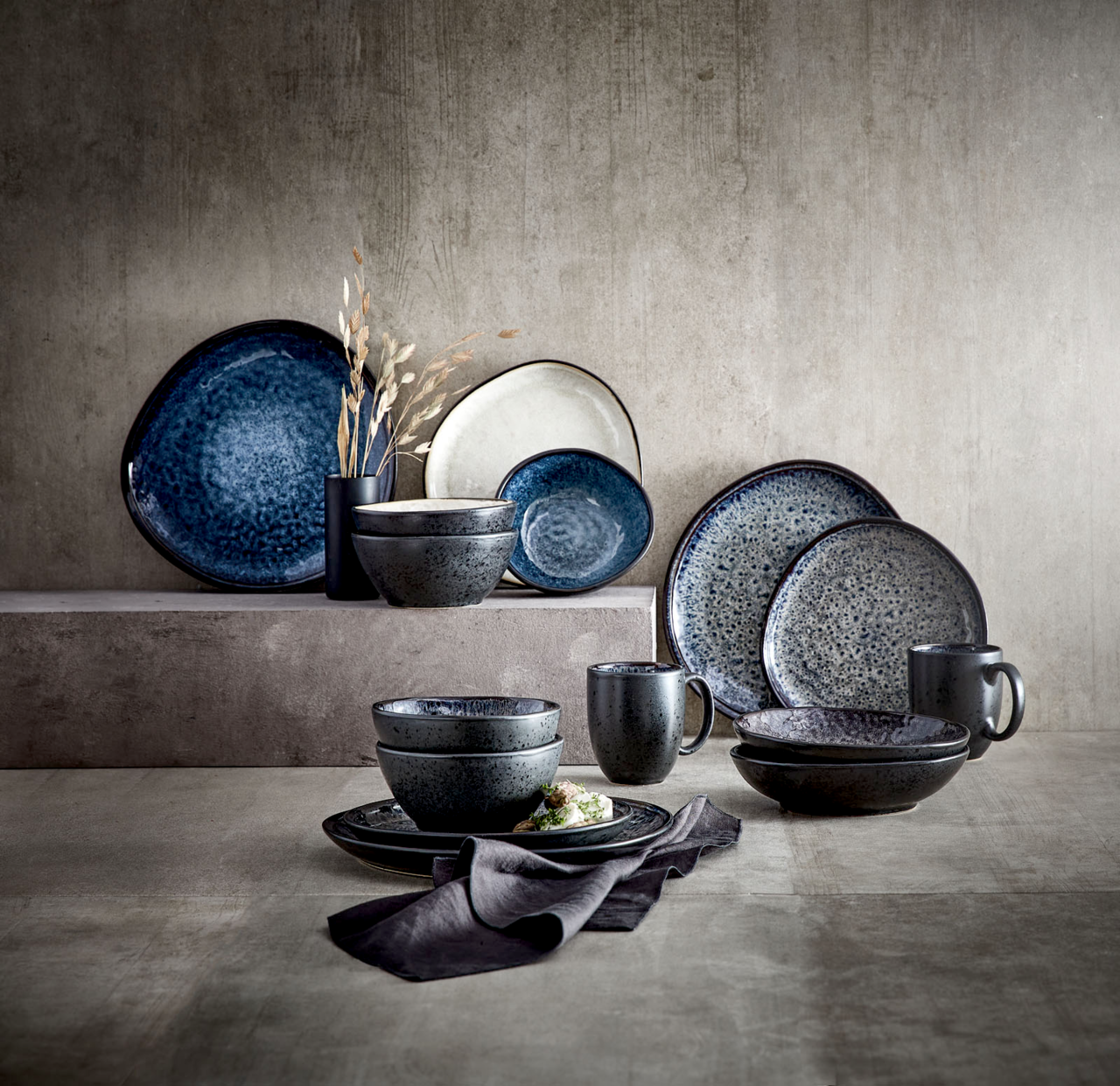 Kombiservice Sören in verschiedenen Blautönen, bestehend aus Tellern, Schalen und Tassen, auf einer strukturierten Oberfläche arrangiert. Seitliche Perspektive.