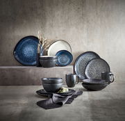 Kombiservice Sören in verschiedenen Blautönen, bestehend aus Tellern, Schalen und Tassen, auf einer strukturierten Oberfläche arrangiert. Seitliche Perspektive.