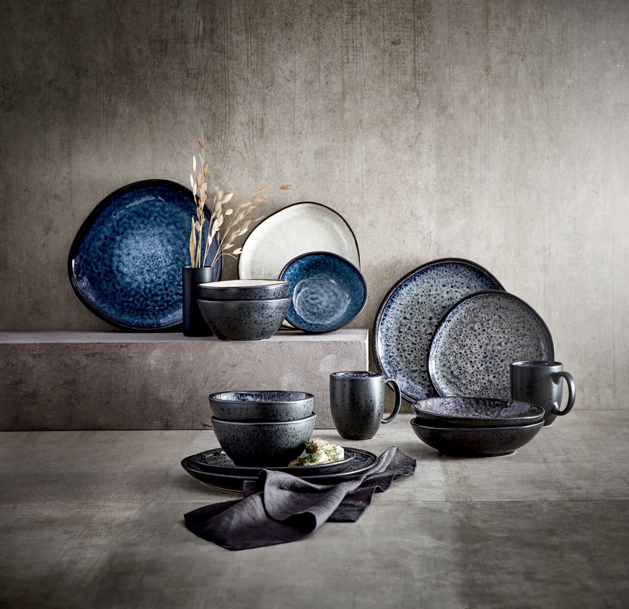 Servies van Sören in verschillende blauwtinten, bestaande uit borden, schalen en kopjes, gerangschikt op een gestructureerd oppervlak. Zijaanzicht.
