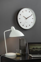Moderne Wanduhr mit weißem Zifferblatt und braunen Zeigern, im Hintergrund eine graue Wand. Im Vordergrund eine weiße Tischlampe, ein geriffelter Glaskrug und ein Laptop auf einem schwarzen Tisch.