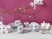 Weißes Porzellan-Kaffeeservice mit rosa Blütenmotiv, Tassen und Untertassen auf hellem Leinentuch vor pinkem Hintergrund, dekoriert mit Magnolienzweigen; Perspektive: leicht von vorne in Augenhöhe.