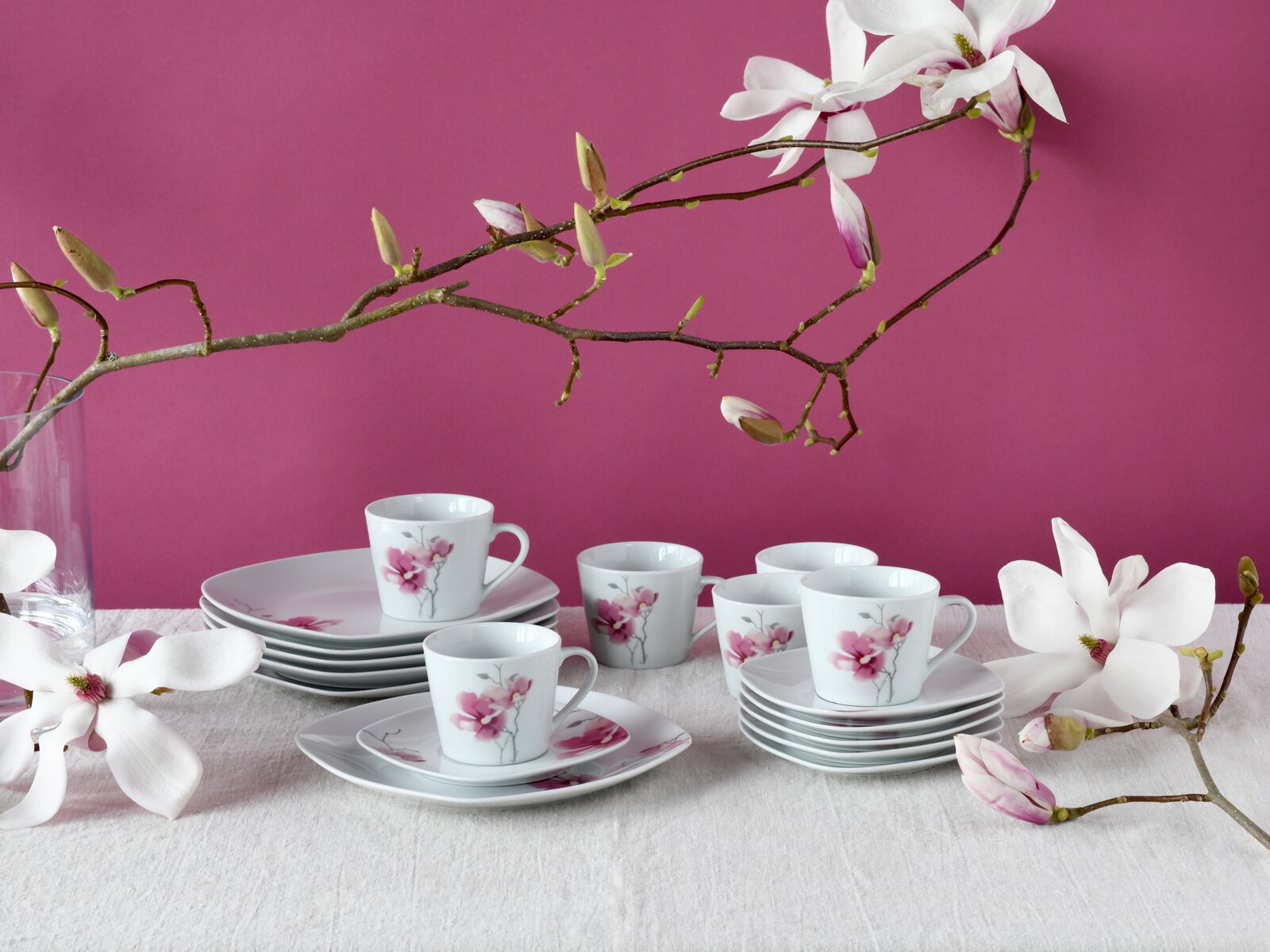 Weißes Porzellan-Kaffeeservice mit rosa Blütenmotiv, Tassen und Untertassen auf hellem Leinentuch vor pinkem Hintergrund, dekoriert mit Magnolienzweigen; Perspektive: leicht von vorne in Augenhöhe.