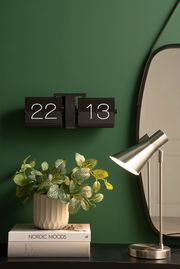 Wanduhr Flip No Case an einer grünen Wand, frontal betrachtet, neben einem Spiegel, einer Tischlampe und einem Pflanzenarrangement auf Büchern.
