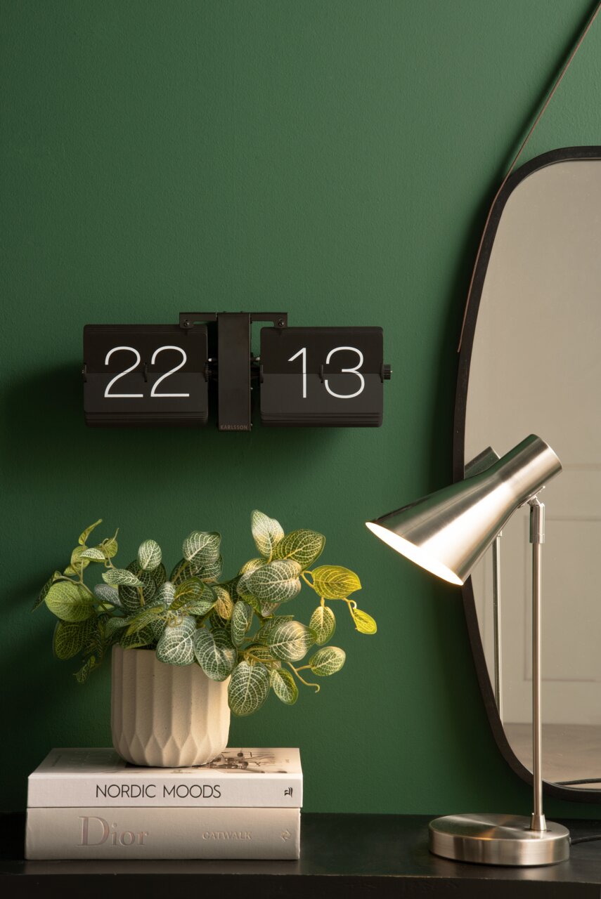 Wandklok Flip No Case aan een groene muur, gezien vanaf de voorkant, naast een spiegel, een tafellamp en een plantenarrangement op boeken.