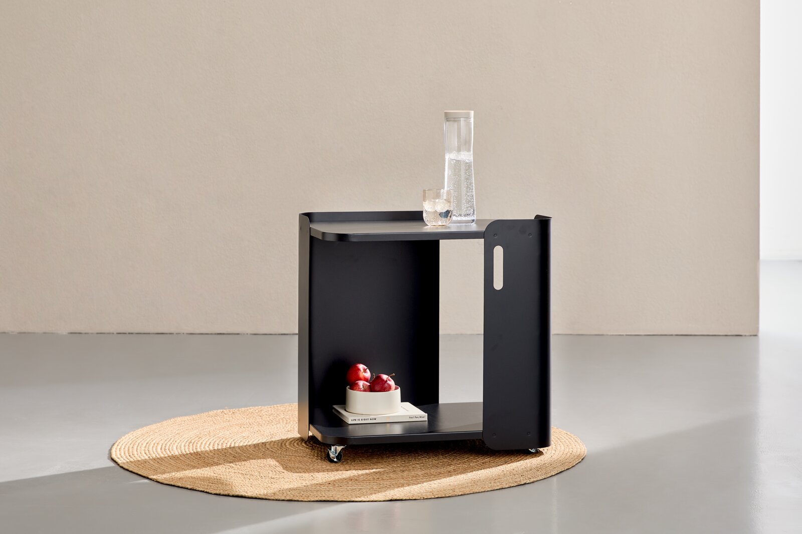 Schwarzer Servierwagen/Beistelltisch aus Metall mit zwei Ablagen und Rollen, auf der oberen Ablage Karaffe und Glas, auf der unteren Ablage Buch und Schale mit roten Früchten; Ansicht schräg von vorne in einem minimalistischen Raum.
