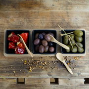 Draufsicht auf ein Servierset aus Bambus mit drei schwarzen Schalen, gefüllt mit gefüllten Paprikaschoten, Oliven und Kapern, auf einem rustikalen Holztisch.