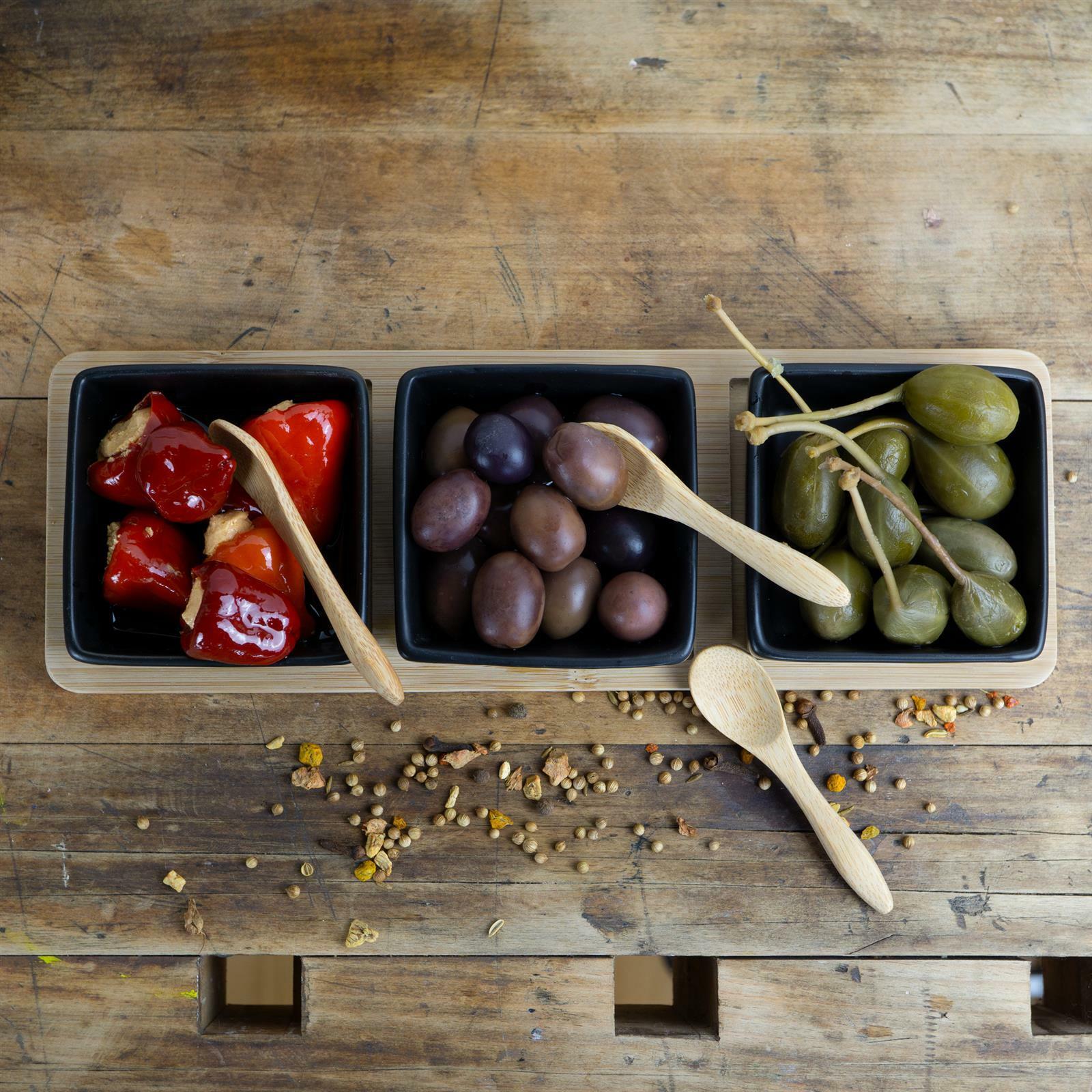 Draufsicht auf ein Servierset aus Bambus mit drei schwarzen Schalen, gefüllt mit gefüllten Paprikaschoten, Oliven und Kapern, auf einem rustikalen Holztisch.