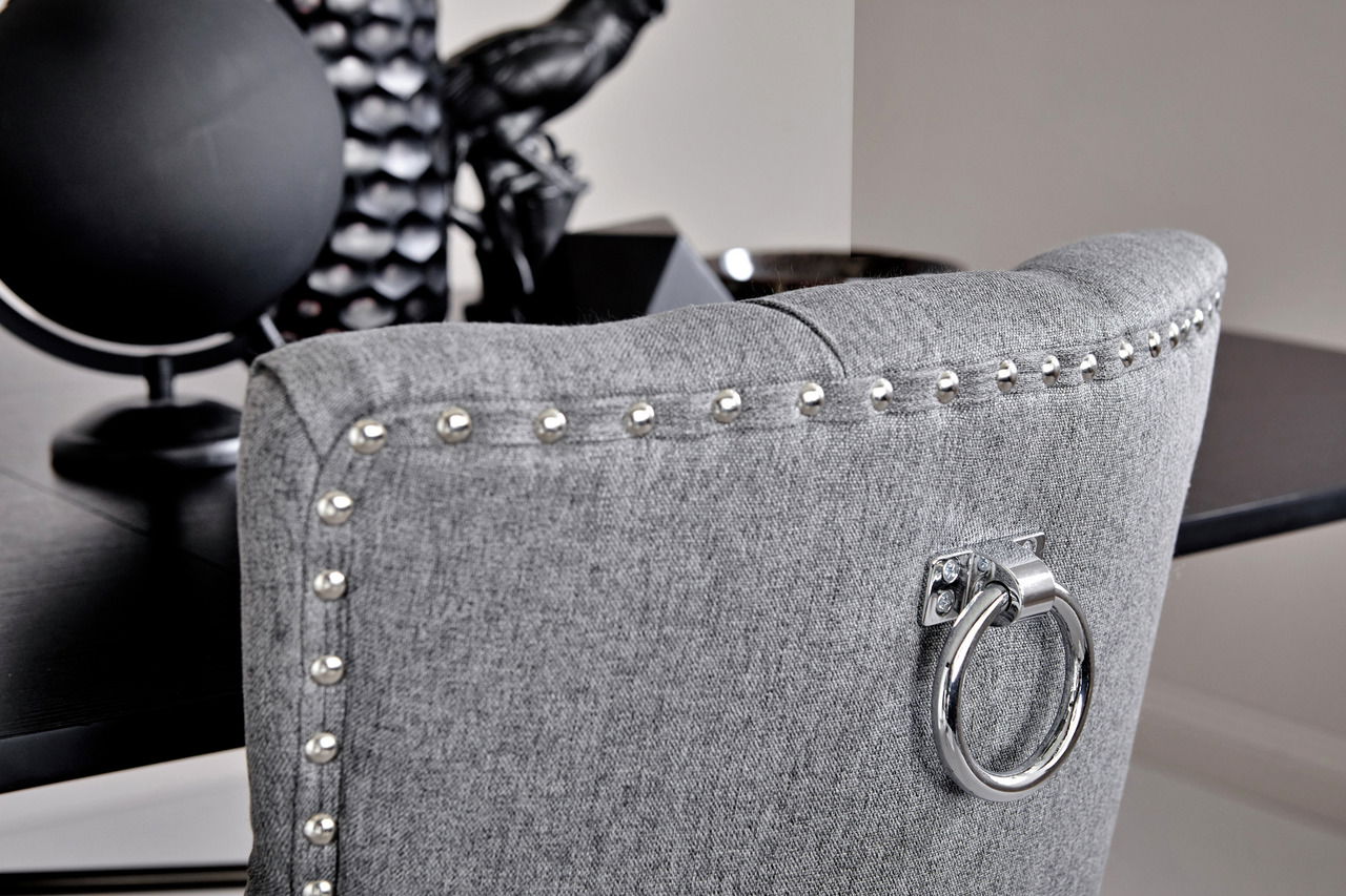 Rückansicht eines grauen Stuhls mit Nietenverzierung und einem dekorativen Metallring an der Rückenlehne, im Hintergrund ein schwarzer Tisch.