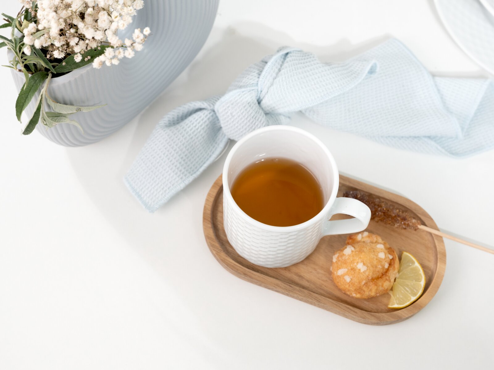 Draufsicht: Weißer Becher mit Tee auf ovalem Holztablett, daneben Zitronenscheibe, kleiner Keks und Kandiszucker-Stick auf hellem Hintergrund mit blauem Tuch und Blumenvase.