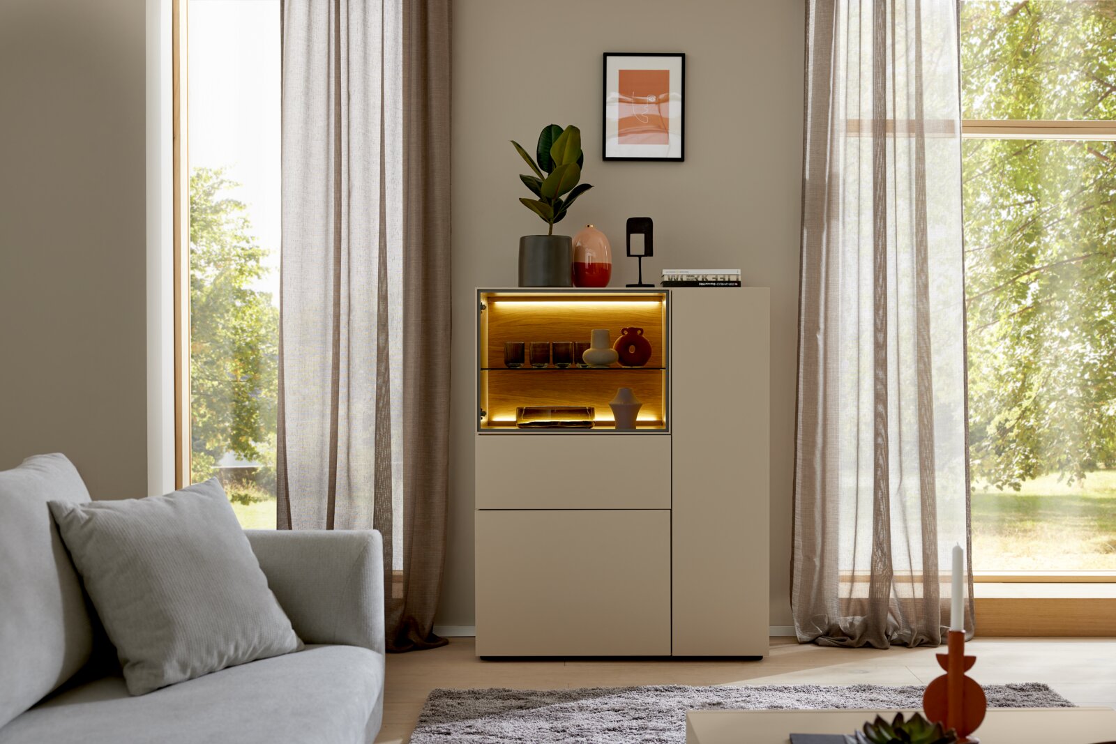 Frontansicht eines modernen beigefarbenen Highboards mit beleuchteter Glasvitrine und dekorativen Wohnaccessoires in einem hellen Wohnzimmer.