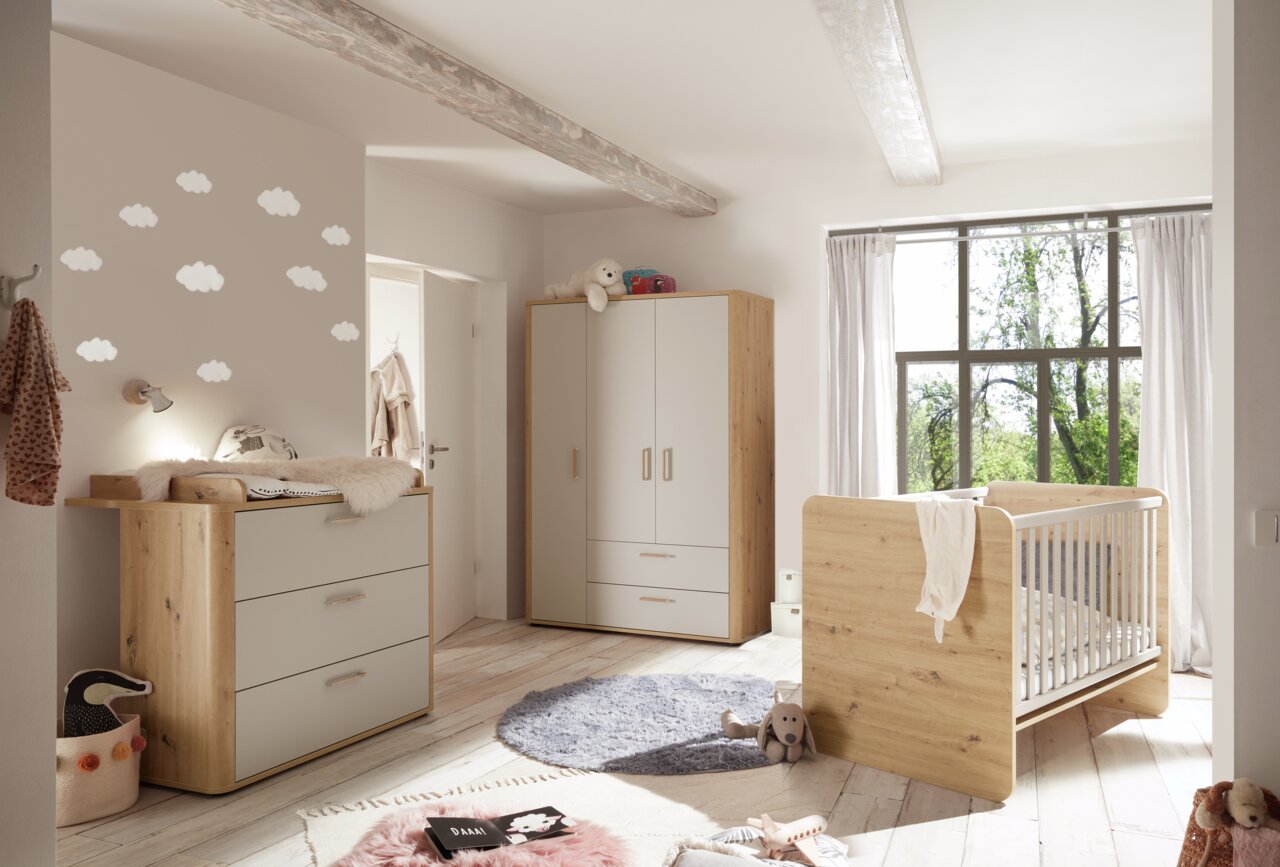 Gemütliches Babyzimmer mit Holzmöbeln, bestehend aus einem Kleiderschrank, einer Wickelkommode und einem Babybett, aus der Perspektive von der Zimmertür aus gesehen.
