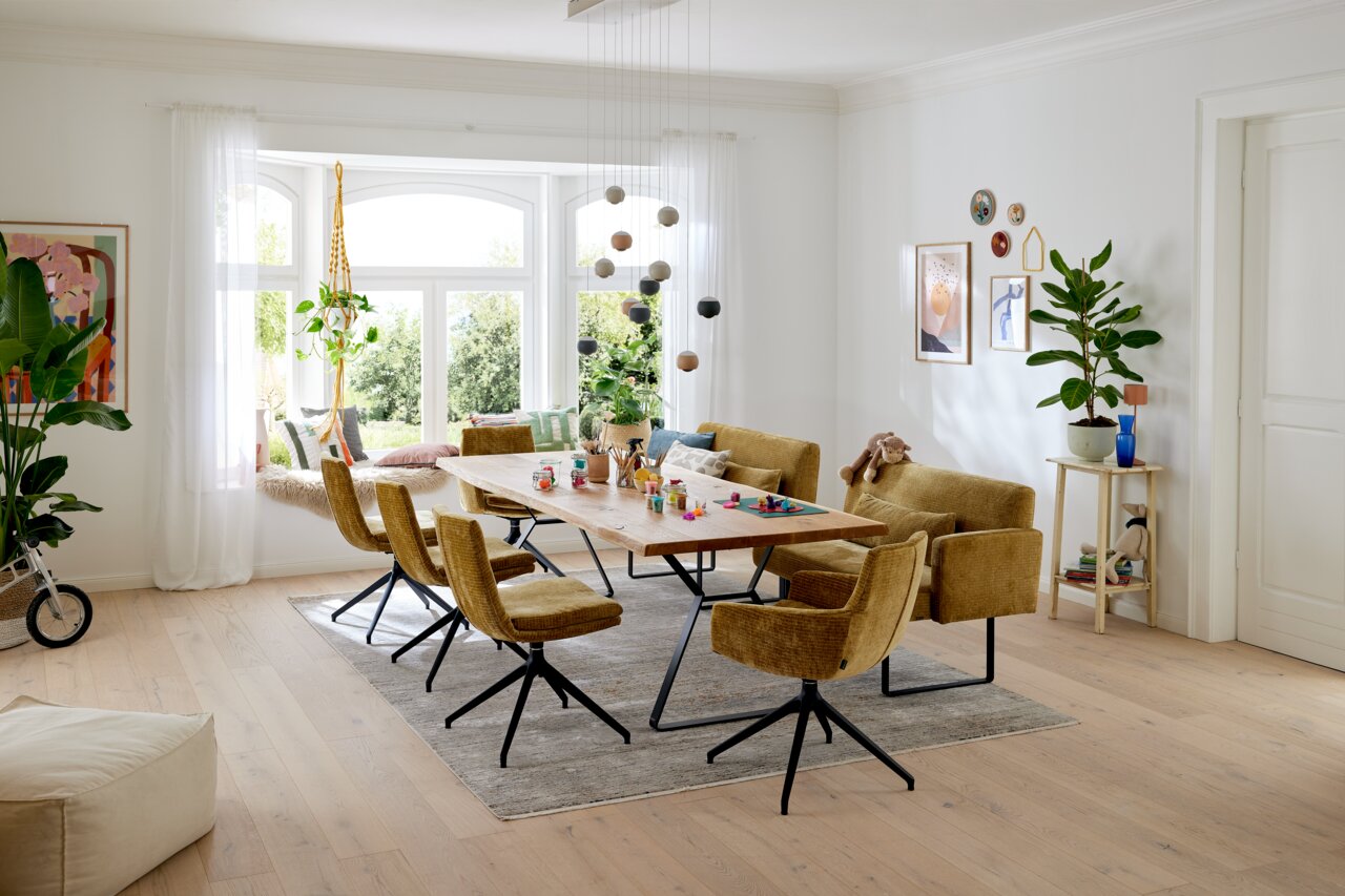 Musterring Eettafel MR T4010 Een moderne eettafel in een lichte woonkamer, omringd door zes gele stoelen, met uitzicht op een groot raam en een groen uitzicht.