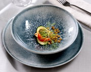 Nahaufnahme eines stilvollen, blauen Tellersets mit 18 Teilen, präsentiert mit einer eleganten Speiseanrichtung aus Linsen, Tomate und Gurke.