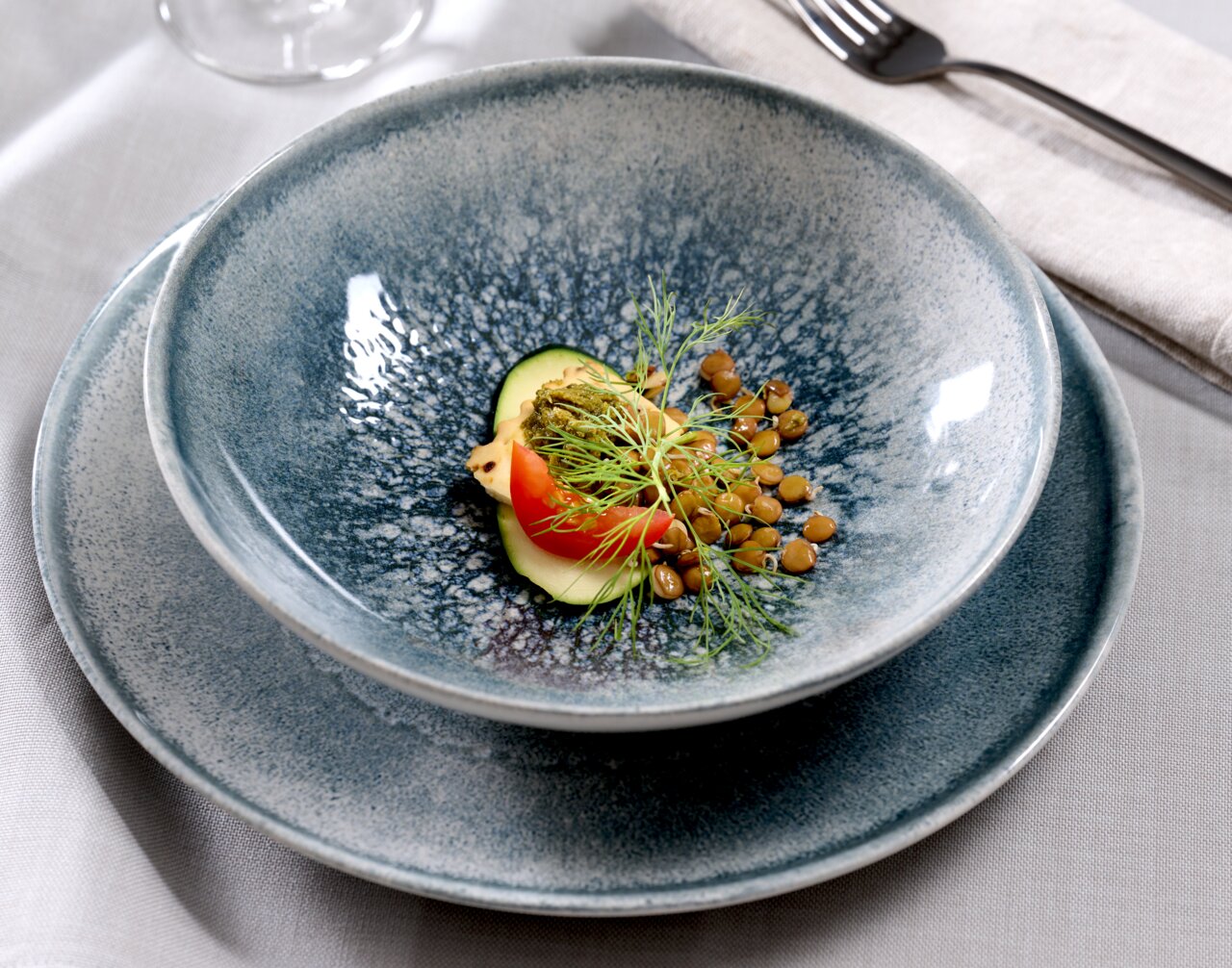 Nahaufnahme eines stilvollen, blauen Tellersets mit 18 Teilen, präsentiert mit einer eleganten Speiseanrichtung aus Linsen, Tomate und Gurke.