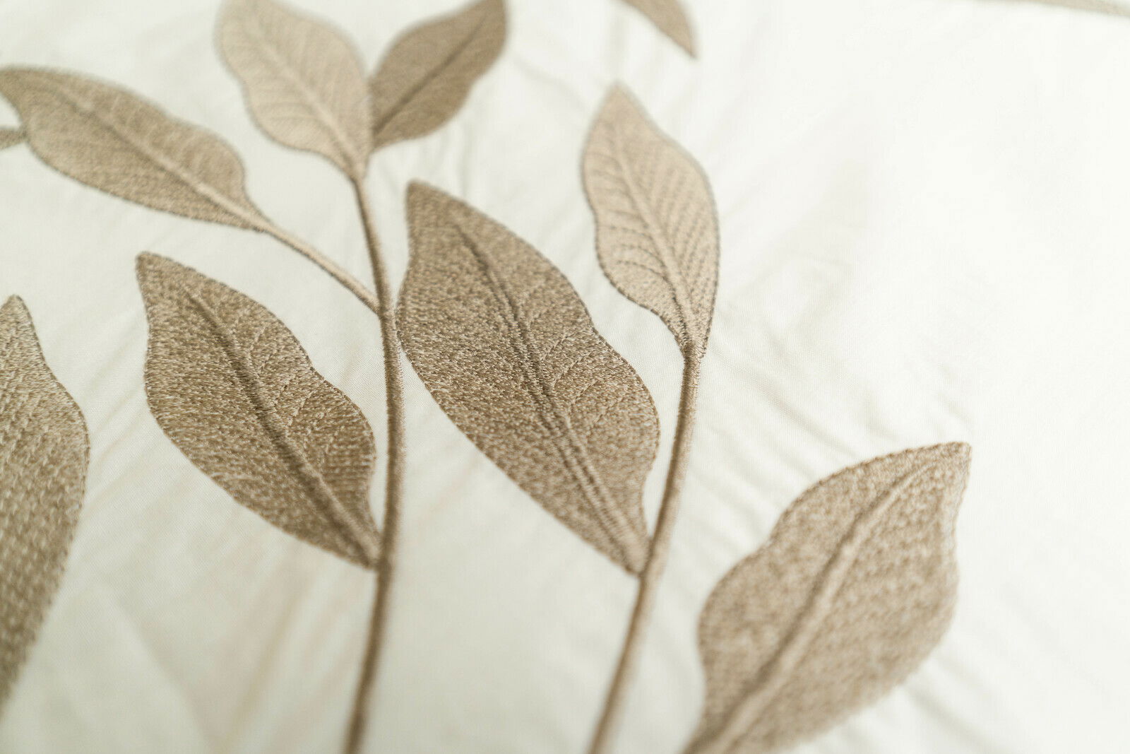 Nahaufnahme von Bettwäsche mit gesticktem Blattmuster in Beige auf weißem Hintergrund, von oben betrachtet.