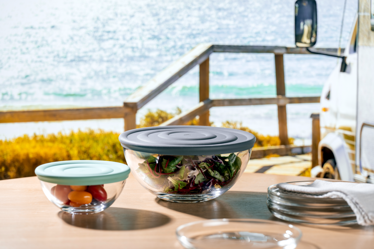 Zwei Glasschalen mit farbigen Deckeln, gefüllt mit Salat und Tomaten, auf einem Tisch mit Meerblick im Hintergrund, aus seitlicher Perspektive.