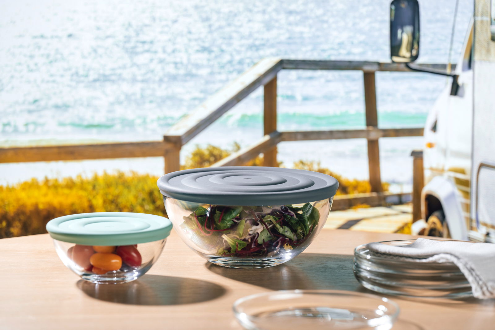 Zwei Glasschalen mit farbigen Deckeln, gefüllt mit Salat und Tomaten, auf einem Tisch mit Meerblick im Hintergrund, aus seitlicher Perspektive.