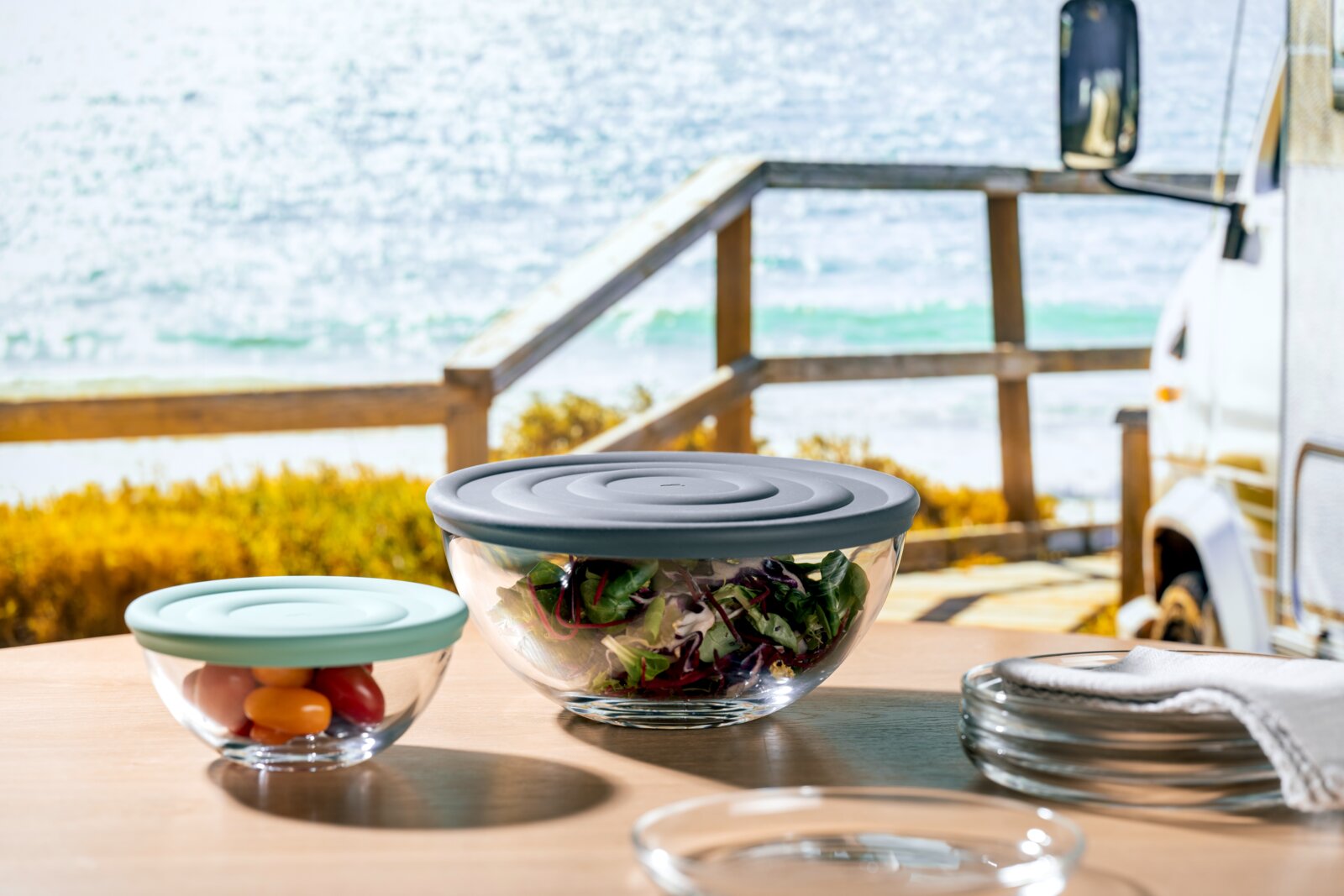 Leonardo Schalen/Deckel 6tlg CUCINA Zwei Glasschalen mit farbigen Deckeln, gefüllt mit Salat und Tomaten, auf einem Tisch mit Meerblick im Hintergrund, aus seitlicher Perspektive.