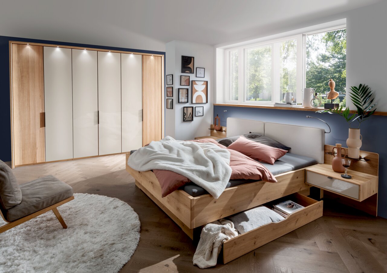 Gemütliches Schlafzimmer mit einem großen Holzbett, das über einen geräumigen Bettschubkasten verfügt. Der Raum ist modern eingerichtet mit einem großen Kleiderschrank und dekorativen Elementen. Perspektive von der Seite des Bettes.