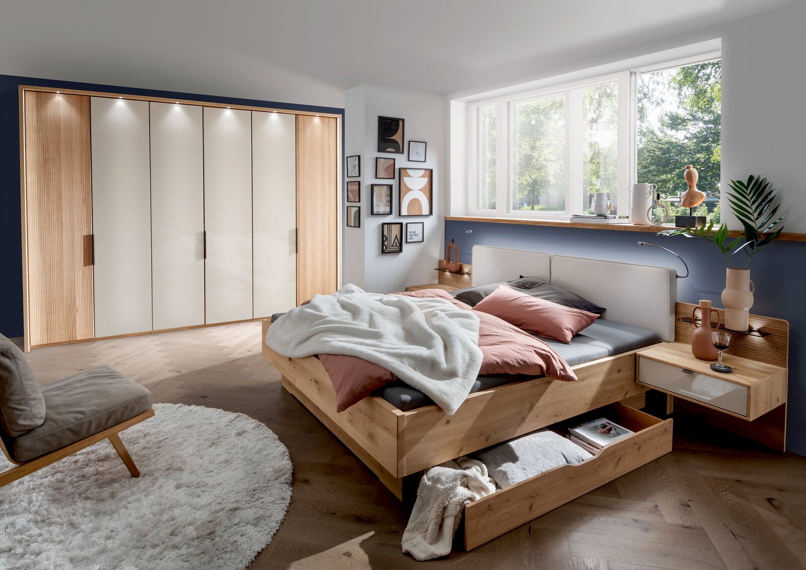 Gemütliches Schlafzimmer mit einem großen Holzbett, das über einen geräumigen Bettschubkasten verfügt. Der Raum ist modern eingerichtet mit einem großen Kleiderschrank und dekorativen Elementen. Perspektive von der Seite des Bettes.