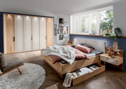 Gemütliches Schlafzimmer mit einem großen Holzbett, das über einen geräumigen Bettschubkasten verfügt. Der Raum ist modern eingerichtet mit einem großen Kleiderschrank und dekorativen Elementen. Perspektive von der Seite des Bettes.