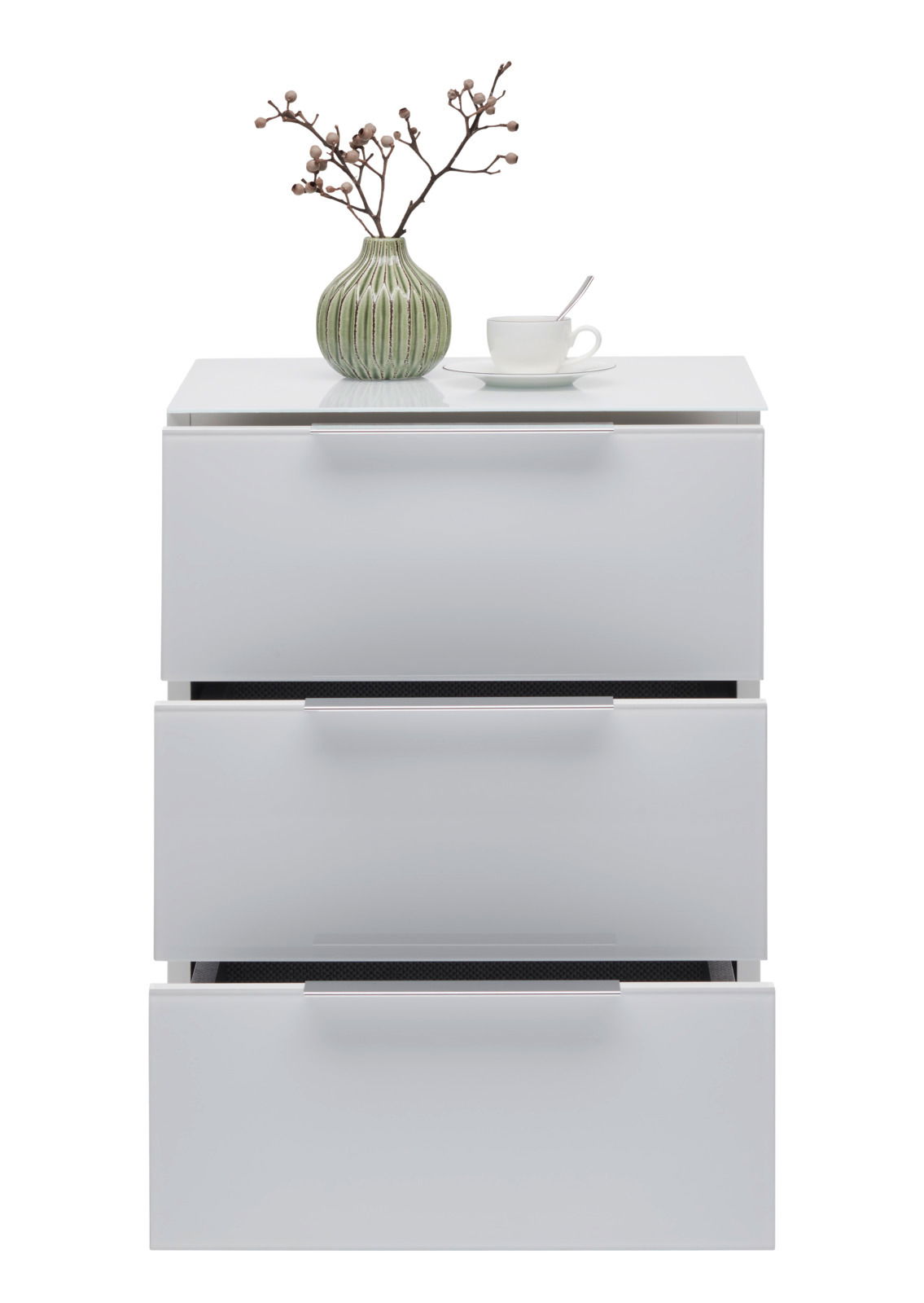 Weiße Nachtkonsole mit drei Schubladen, darauf eine grüne Vase mit Zweigen und eine weiße Tasse mit Löffel, frontale Ansicht.