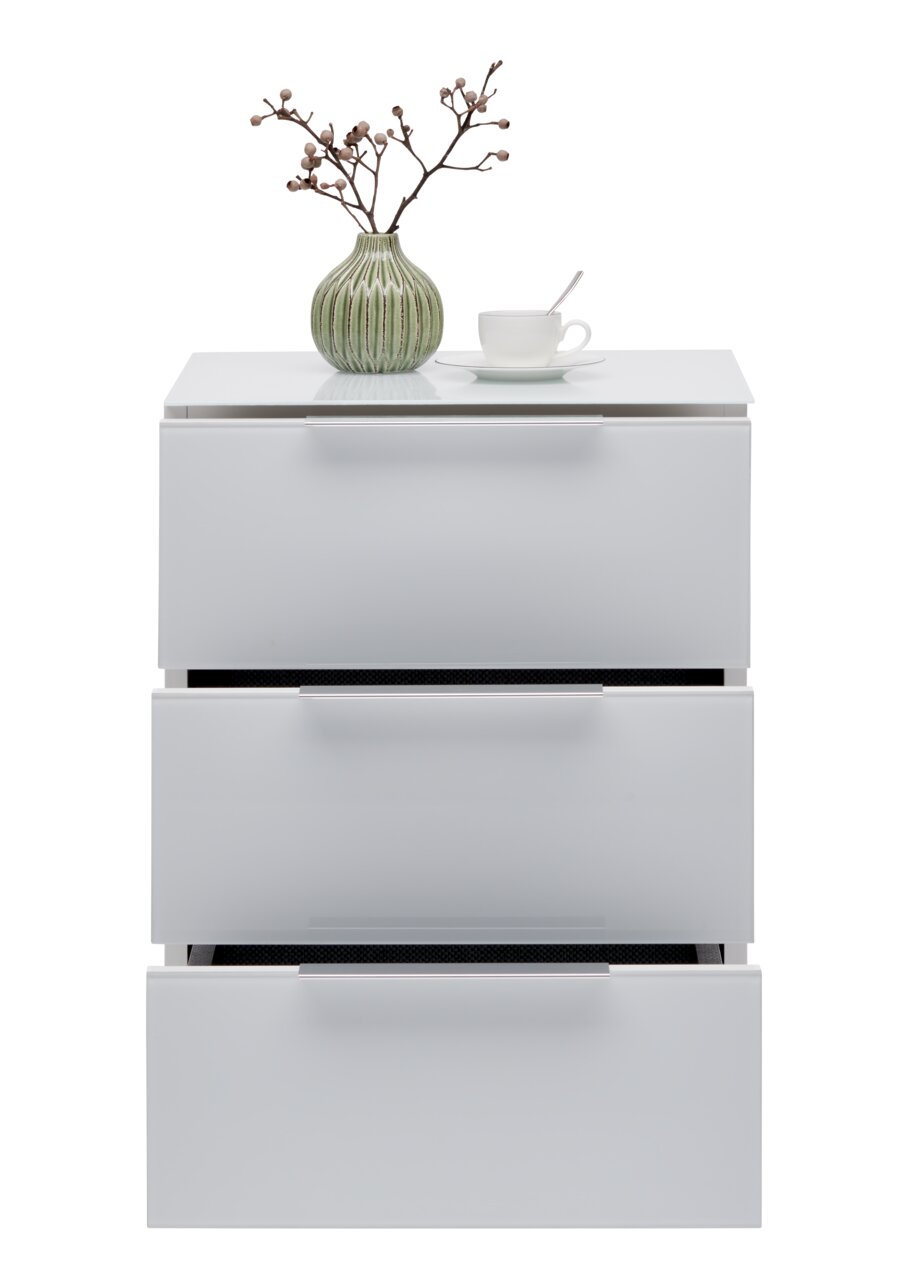Weiße Nachtkonsole mit drei Schubladen, darauf eine grüne Vase mit Zweigen und eine weiße Tasse mit Löffel, frontale Ansicht.