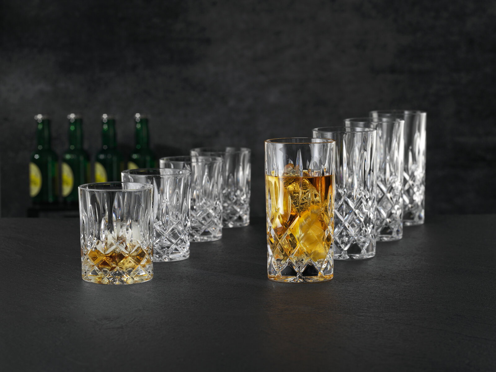 Kristallglas-Whiskygläser und Longdrinkgläser mit Facettenschliff auf dunkler Oberfläche, ein Glas mit Whisky und Eis im Vordergrund, perspektivische Schrägansicht von vorn