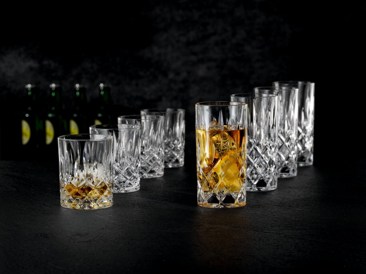 Nachtmann Becherset 8-tlg. NOBLESSE Whiskyglazen en longdrinkglazen van kristalglas met facetten op een donkere achtergrond, een glas met whisky en ijs op de voorgrond, een perspectivisch schuin aanzicht van voren