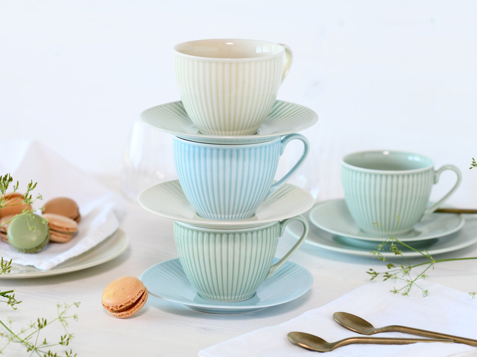 Stapel aus geriffelten Porzellan-Kaffeetassen mit passenden Untertassen in Pastellfarben (Creme, Hellblau, Mint) auf gedecktem Tisch mit Macarons und Löffeln, frontale Ansicht in leichter Schrägperspektive
