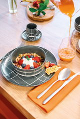 Frühstückstisch mit einem flachen Teller von 27 cm Durchmesser, darauf eine Schale mit Müsli, frischen Beeren und Joghurt, daneben ein Glas Orangensaft und Besteck auf einer orangefarbenen Serviette. Perspektive von oben.
