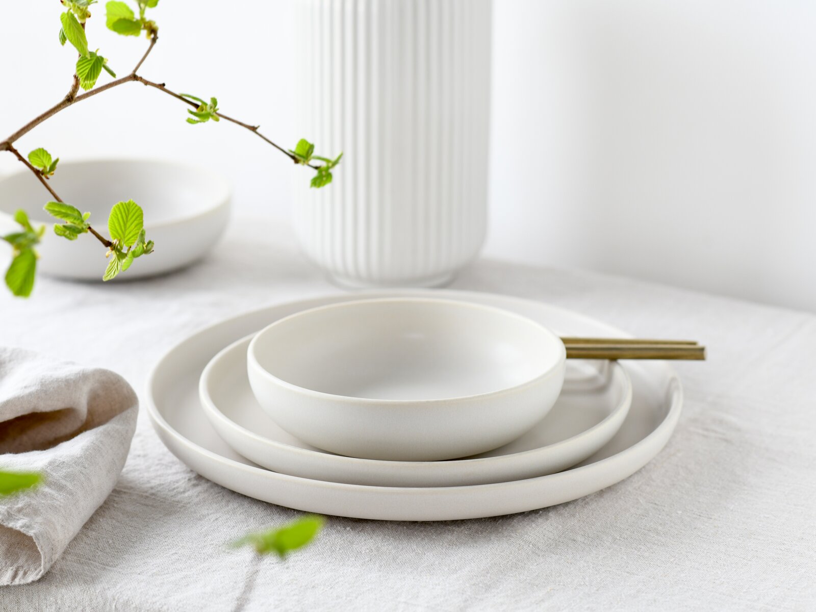 Minimalistisches weißes Geschirr-Set mit Schale auf zwei Tellern und goldfarbenen Stäbchen auf hellem Leinentuch, seitliche Perspektive leicht von oben; im Hintergrund eine geriffelte weiße Vase und ein Zweig mit frischen grünen Blättern.