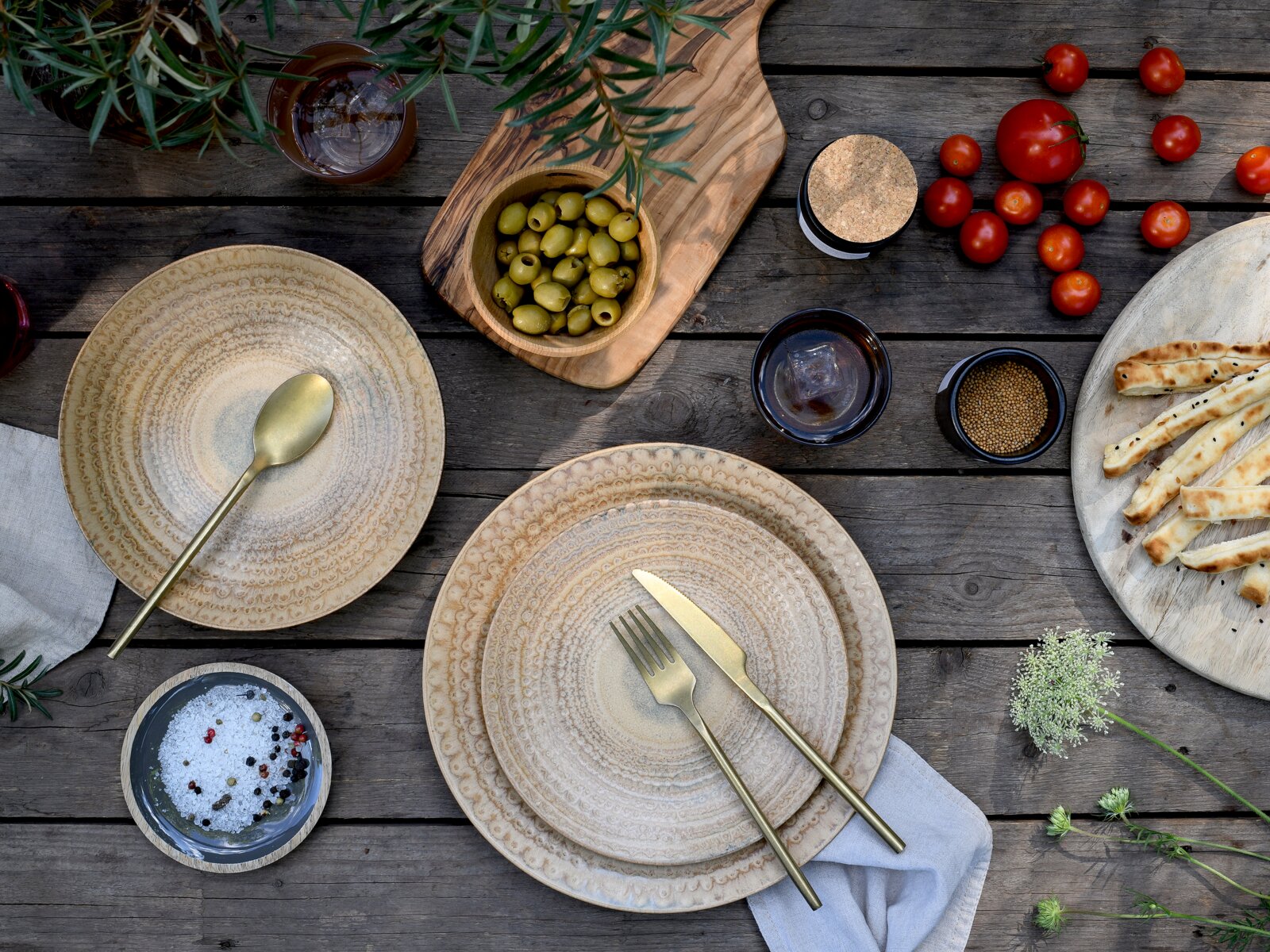Draufsicht auf ein rustikales Tisch-Arrangement mit beigen Keramiktellern, goldfarbenem Besteck, grünen Oliven in Holzschale, Kirschtomaten, Brotstangen und Gewürzen auf verwittertem Holztisch.