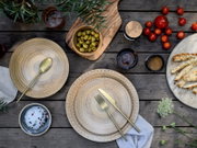 Draufsicht auf ein rustikales Tisch-Arrangement mit beigen Keramiktellern, goldfarbenem Besteck, grünen Oliven in Holzschale, Kirschtomaten, Brotstangen und Gewürzen auf verwittertem Holztisch.