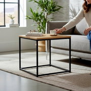 Moderner Beistelltisch mit Holzplatte und schwarzem Metallgestell neben einem grauen Sofa, darauf Bücher und eine Tasse; Perspektive: leicht schräg von vorne auf Tischhöhe.