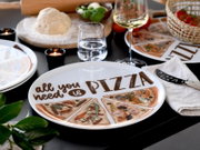 Schräg von oben fotografierter weißer Pizzateller mit Aufschrift „all you need is PIZZA“ und illustrierten Pizzastücken, auf gedecktem Tisch mit Weinglas, Wasserglas, Kerzen und Zutaten im Hintergrund.