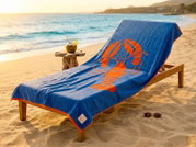 Blaue Stranddecke/Badetuch mit großem orangefarbenem Hummer-Motiv und orangefarbener Umrandung auf einer Holzliege am Sandstrand, seitliche Perspektive leicht von vorn; daneben kleiner Holztisch mit Kokosdrink und Sonnenbrille im Vordergrund, Meer im Hint