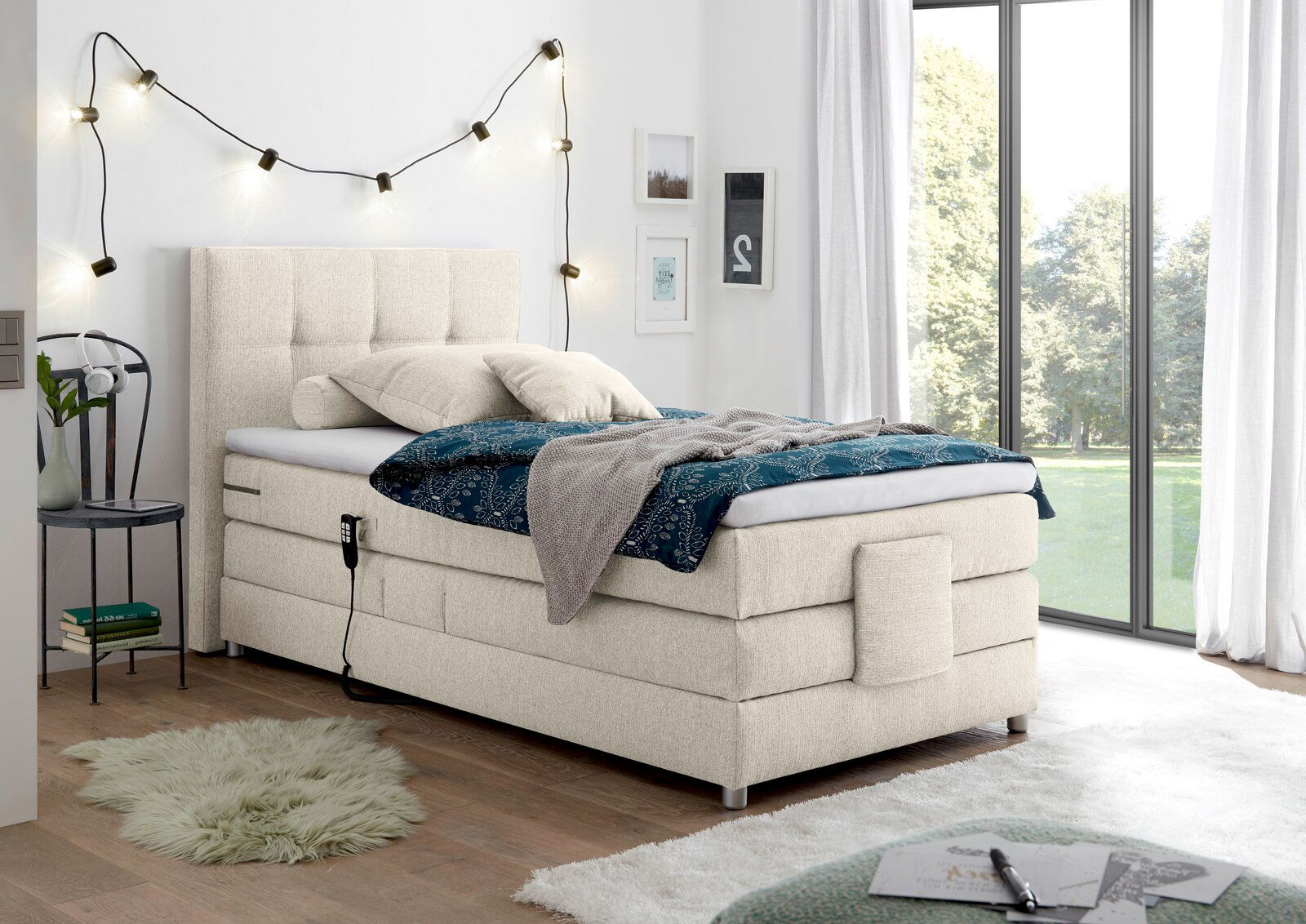 Beiges Boxspringbett in einem modernen Schlafzimmer, seitliche Perspektive mit Dekoration und großen Fenstern im Hintergrund.