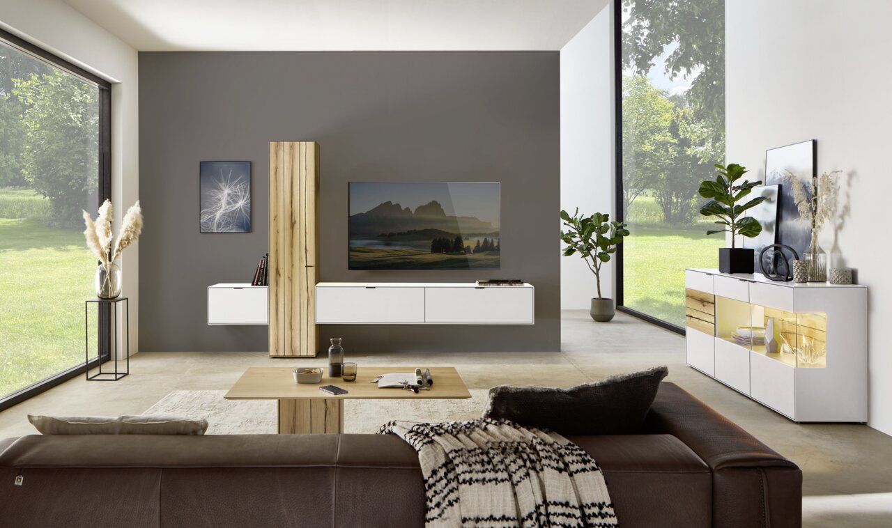 Moderne Wohnzimmeransicht mit Wandkombination, bestehend aus weißen Schränken und Holzelementen, im Vordergrund ein braunes Sofa, seitliche Perspektive.