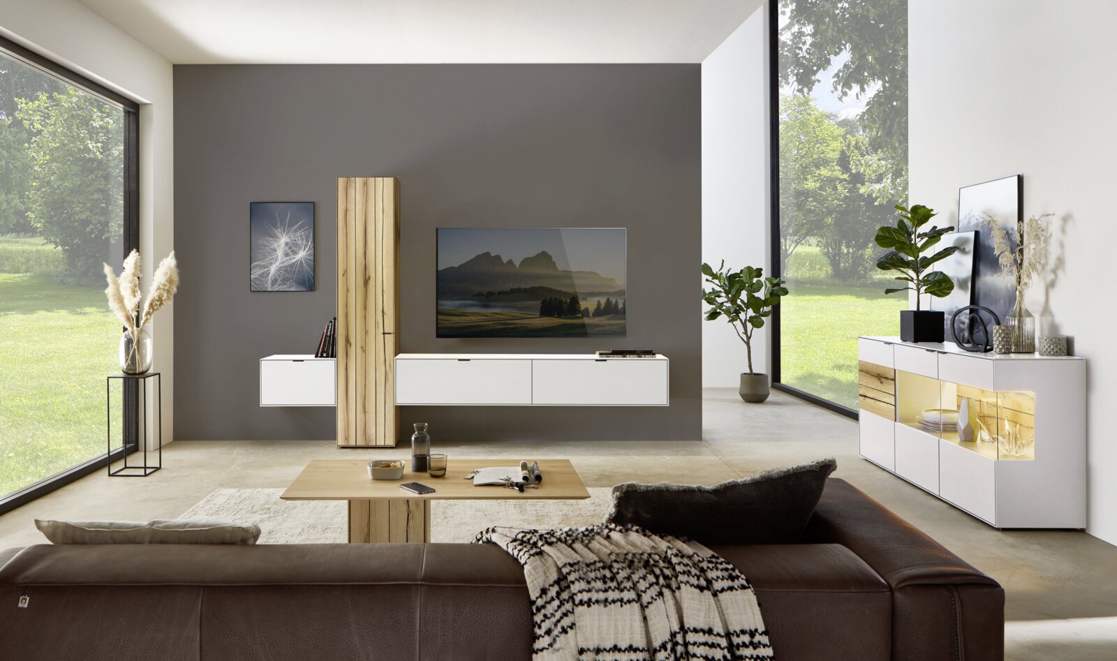 Moderne Wohnzimmeransicht mit Wandkombination, bestehend aus weißen Schränken und Holzelementen, im Vordergrund ein braunes Sofa, seitliche Perspektive.