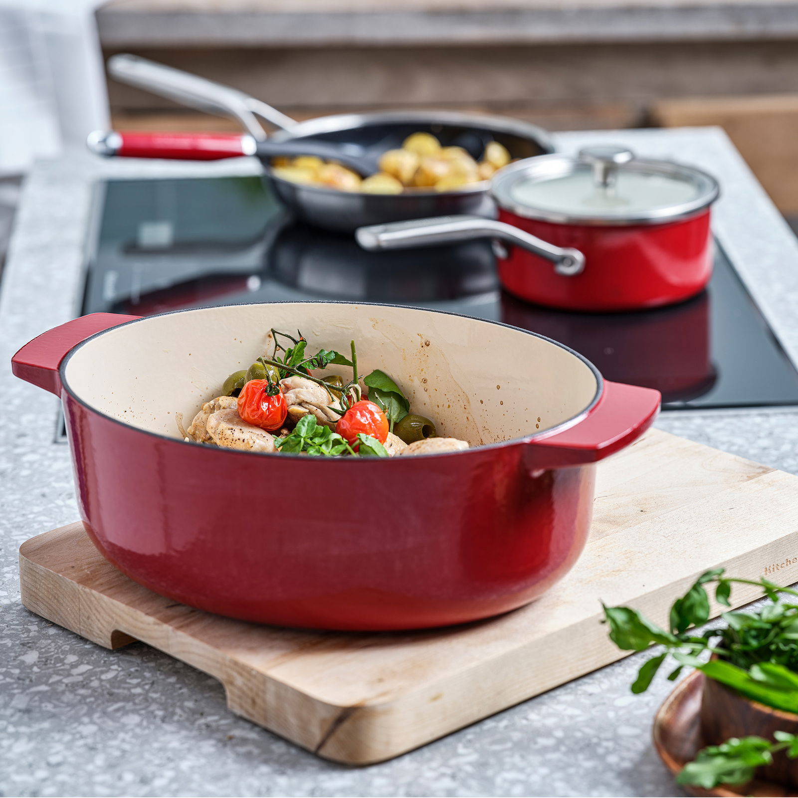 Roter ovaler Gusseisen-Bräter mit Griffen, gefüllt mit frischen Zutaten, auf einem Holzbrett in einer modernen Küche, seitliche Perspektive.