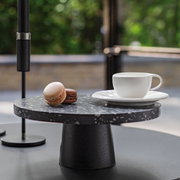 Weiße Kaffeetasse NewMoon 180ml auf einem Unterteller, seitliche Perspektive, neben zwei Macarons auf einem schwarzen, runden Podest.
