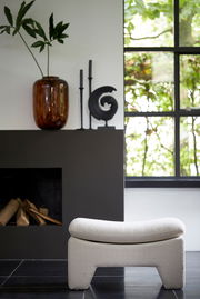 Moderner Sitzpuff in heller Farbe, frontal vor einem Kamin und einem Fenster mit Blick ins Grüne.