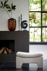 Moderner Sitzpuff in heller Farbe, frontal vor einem Kamin und einem Fenster mit Blick ins Grüne.